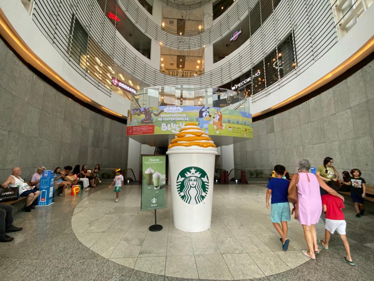 Copo gigante Starbucks Frappuccino no átrio de shopping center com campanha Pistacho e banner do Playground da Bluey ao fundo